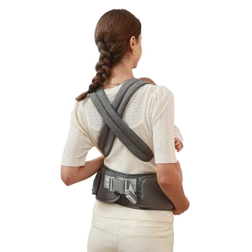 Woman wearing ergonomic baby carrier with crossed shoulder straps and hip seat in grey color